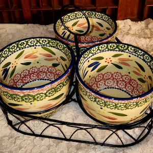 May include: A set of three ceramic bowls with a floral design, held in a black metal serving tray. The bowls have a cream-colored base with blue, green, pink, and yellow floral patterns. The rims are trimmed with a blue border.
