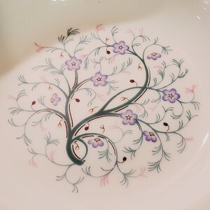 May include: A white ceramic plate with a delicate floral design. The design features a stylised tree with purple flowers and green leaves.