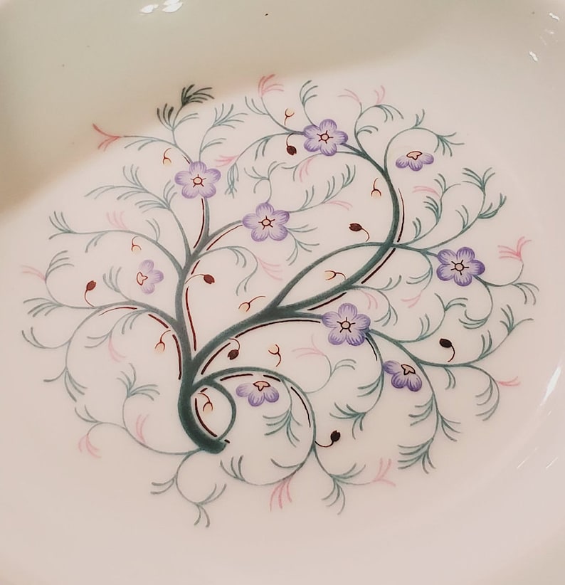 May include: A white ceramic plate with a delicate floral design. The design features a stylised tree with purple flowers and green leaves.