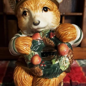 May include: A ceramic figurine of a brown bear wearing a green and red hat and holding a green wreath with red berries and pine cones.