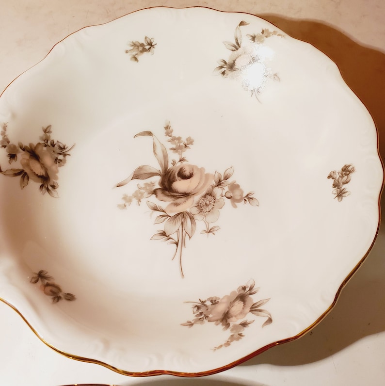 May include: A white porcelain bowl with a scalloped rim and a gold trim. The bowl features a floral design with brown roses and leaves.
