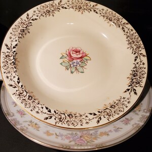 May include: A white ceramic soup bowl with a gold rim and a black floral design. The bowl features a pink rose with green leaves and a gold border.