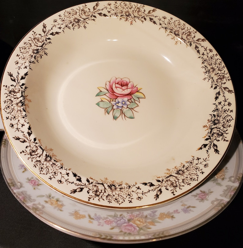 May include: A white ceramic soup bowl with a gold rim and a black floral design. The bowl features a pink rose with green leaves and a gold border.