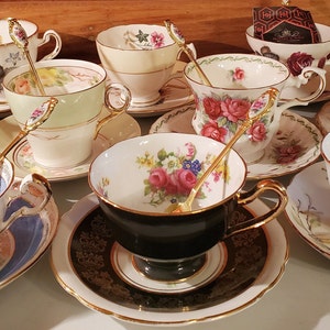 May include: A collection of vintage teacups and saucers with gold trim, each with a unique floral design. Gold spoons rest in the cups. Some cups have tea bags. The teacups are on a light-colored surface.