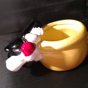 Op de afbeelding: Een gele keramische pot in de vorm van een pot met een zwarte kat die aan de ene kant uitkijkt en een gele vogel die aan de andere kant uitkijkt.