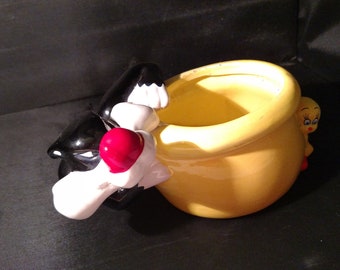 Puede incluir: Un macetero de cerámica amarillo con forma de olla con un gato negro asomándose por un lado y un pájaro amarillo asomándose por el otro lado.