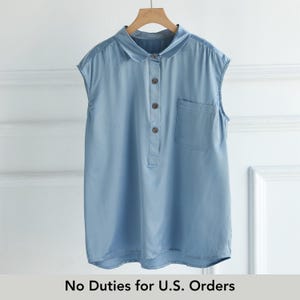 May include: A sleeveless light blue collared shirt featuring a partial button-down front with four brown buttons and a single chest pocket on the left side. The garment is displayed on a wooden hanger against a white paneled wall.