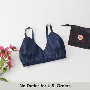 Könnte beinhalten: Ein marineblaues Seiden-Bralette mit verstellbaren Trägern und gerafftem Design. Ein kleiner schwarzer Kordelzugbeutel mit Logo befindet sich neben dem Bralette. Rosa Blumen befinden sich auf der linken Seite des Bildes. Der Text "No Duties for U.S. Orders" steht unten.