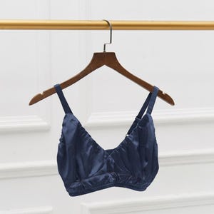 May include: A navy blue bralette with adjustable straps hangs on a wooden hanger. The bralette is made of a silky material and has a V-neck design. The garment is displayed against a neutral background.
