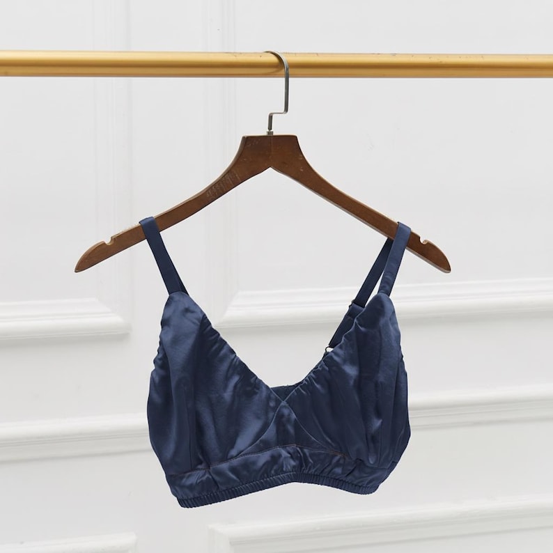 May include: A navy blue bralette with adjustable straps hangs on a wooden hanger. The bralette is made of a silky material and has a V-neck design. The garment is displayed against a neutral background.