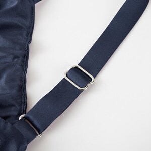 May include: Close-up of a navy blue satin strap with a silver-colored metal buckle. The strap is angled across the frame, with a portion of the garment visible on the left side. The background is plain white.