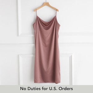 Könnte beinhalten: Ein staubrosafarbenes Slip-Kleid mit Spaghettiträgern hängt an einem Holzbügel. Das Kleid hat einen Wasserfallausschnitt und eine fließende, seidige Optik. Der Text "No Duties for U.S. Orders" ist unten im Bild zu sehen.