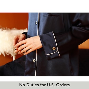 Pode incluir: Close-up de um top de pijama azul marinho com debrum e botões brancos. As mangas compridas têm punhos abotoados. O texto "No Duties for U.S. Orders" está na parte inferior da imagem.