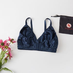 May include: A navy blue silk bra with adjustable straps and a hook-and-eye closure. The bra is displayed on a white surface next to a small black bag with the text "Soft Strokes Silk" and a pink flower arrangement.