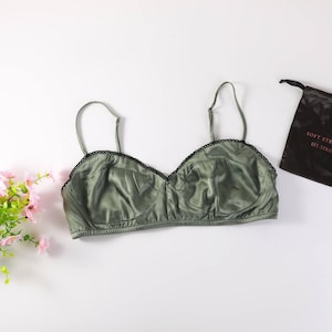 Può includere: Un bralette verde oliva con una finitura liscia e satinata, dotato di sottili spalline regolabili e di un delicato bordo in pizzo nero smerlato lungo la parte superiore. Il capo presenta coppe morbide e una fascia elastica inferiore.