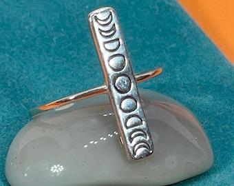 May include: A silver ring featuring a vertical bar with a series of moon phase designs. The ring is set on a smooth, gray stone against a teal background. The ring's design is detailed and the metal has a polished finish.