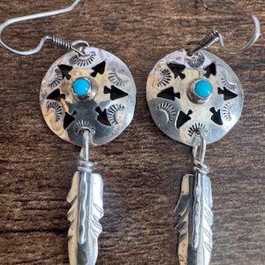 May include: A pair of silver dangle earrings featuring a circular disc with cut-out arrow shapes and stamped patterns. Each disc is centered with a small, round, bright blue turquoise stone and hangs from a silver wire hook with a feather charm.