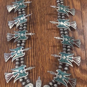 May include: A silver necklace with turquoise and red accents. The necklace features a series of bird-shaped pendants with turquoise inlay, connected by silver beads. The central pendant is a larger bird design.