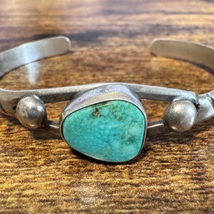 May include: A silver cuff bracelet featuring a central, irregular, teal-colored turquoise stone. The metal band is accented with two smooth, round silver spheres on either side of the stone and includes etched line details along the sides.