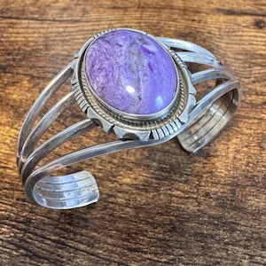 May include: A silver cuff bracelet featuring a large, oval-shaped purple charoite stone set in a decorative bezel. The band consists of four parallel silver wires that merge at the ends, with a notched, textured border surrounding the central stone.