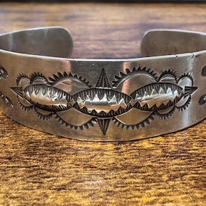 May include: A silver cuff bracelet featuring a wide band with a polished finish. The surface is decorated with a central row of three embossed oval shapes, each surrounded by intricate sunburst stamp patterns and fine serrated edge detailing.