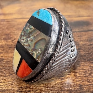 May include: A silver ring with a large, rectangular face. The face is inlaid with turquoise, black, abalone shell, white, and red stones. The ring has a detailed, textured band with a feather design.