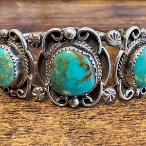 May include: A silver cuff bracelet featuring three oval turquoise stones with brown matrix patterns. The metalwork includes decorative scrollwork, twisted wire borders around each stone, and small circular accents between the settings.
