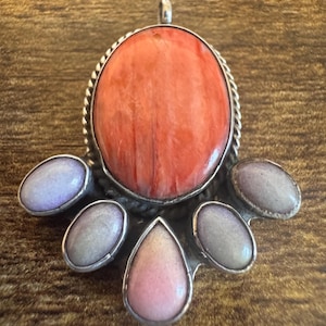 May include: A silver pendant featuring a large, oval, orange-red stone at the center, surrounded by a twisted silver border. Below the central stone are four smaller, smooth, light purple and pink stones set in silver, arranged in a fan shape.