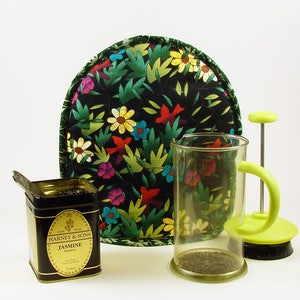 May include: A tea set featuring a black floral tea cozy, a glass French press, and a tin of Harney & Sons Jasmine tea. The cozy has a colorful floral pattern. The French press has a green handle and plunger. The tea tin is black and gold.