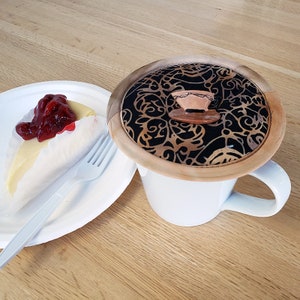 May include: A white ceramic mug with a decorative lid featuring a black and gold pattern and a small teacup accent. A slice of cheesecake with cherry topping and a plastic fork sit on a paper plate.