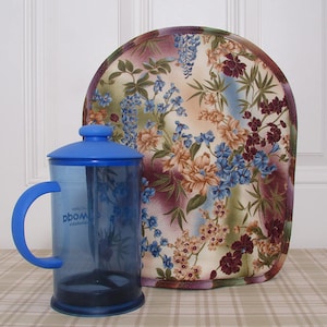 May include: A blue French press coffee maker with a matching lid and handle. The transparent carafe has a visible filter. A floral-patterned tea cozy with blue, purple, and tan flowers is behind the press.