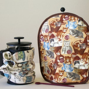 May include: A French press coffee maker with a patterned cozy and a matching tea cozy. The tea cozy features a brown border and a beige background with a repeating pattern of kittens, teacups, and yarn balls. A purple spoon rests in front.