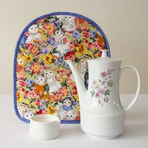 May include: A white porcelain coffee pot with a floral design, a small creamer, and a padded tea cozy featuring a cat and floral pattern. The cozy has a blue border and a colorful floral and cat print.