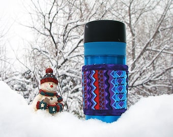 May include: A bright blue travel mug with a black lid, featuring a fabric sleeve decorated with a purple, red, and blue geometric pattern. Beside the mug stands a small snowman figurine wearing a red hat and scarf, holding a tiny brown object.