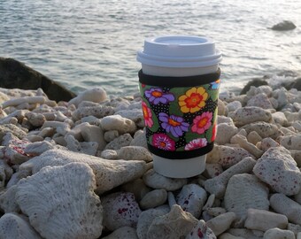 May include: A white disposable coffee cup with a white lid and a colorful floral sleeve. The sleeve features a black background with vibrant flowers in shades of pink, purple, yellow, and orange. The cup is resting on a bed of white rocks near the ocean.