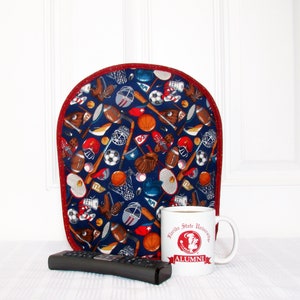 May include: A navy blue padded chair back cover with a red trim, featuring a sports-themed pattern of footballs, baseballs, soccer balls, and hockey sticks. A white mug with a university logo and a black remote control are in front of the chair back.