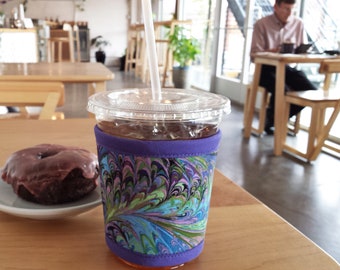 May include: A clear plastic cup filled with iced coffee, featuring a decorative purple and patterned fabric sleeve. A white straw is inserted into the drink. A chocolate glazed donut sits on a white plate. The items are on a wooden table.