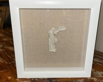 May include: A white square shadow box frame containing a small, off-white sculptural relief of the Winged Victory of Samothrace. The sculpture is centered against a light beige, textured linen fabric backing within the frame's interior.