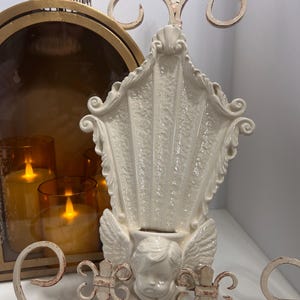 Könnte beinhalten: Eine weiße Keramik-Wandleuchte mit Muscheldesign, Engelsgesicht und Flügeln. Die Leuchte hat eine cremefarbene, antike Oberfläche und dekorative Metallverzierungen. Im Hintergrund eine goldfarbene Laterne und Kerzen.