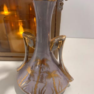 May include: A lavender-colored ceramic vase with gold accents and a palm tree design. The vase has a slender neck and flared handles. The gold detailing includes the rim and handles. The vase is likely for decorative purposes.
