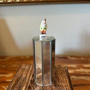 Puede incluir: Una pequeña figura de porcelana del personaje Dormilón de Blancanieves, con una túnica verde claro y pantalones rojos, sobre un pedestal cilíndrico de espejo con el texto DORMEUR y DISNEY impreso en la base de la figura.