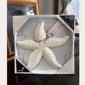 May include: A white starfish wall decor piece with a textured, carved appearance, packaged in a square cardboard frame. The item features a tag labelled T.I. Design and a corner label marked Coastal. The starfish has five arms and a matte finish.