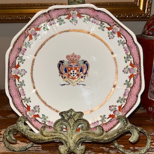 Könnte beinhalten: Ein dekorativer antiker Teller mit einem rosa-weißen Blumenrand und einem Goldrand. Der Teller hat ein zentrales Wappenmotiv und wird auf einem dekorativen Metallständer präsentiert. Der Teller ist ein Vintage-Stück, wahrscheinlich Porzellan.