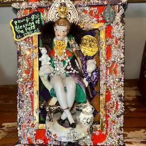 May include: A decorative framed figurine featuring a seated figure in regal attire, holding a scepter and a small white animal. The frame is embellished with sparkling crystals, gold accents, and the text "Paw-di-gras Blessing of the Pets St. Frances".