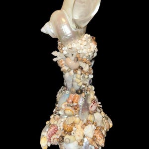 May include: A decorative sculpture shaped like a dress, entirely covered in a dense mosaic of various seashells, including small white spiral shells, tan bivalves, and iridescent fragments, topped with a large, smooth, pearlescent conch shell.