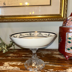 May include: A white and gold footed bowl with a clear, cut-glass base. The bowl has a black floral pattern around the rim. The bowl is on a wooden surface, with a framed picture and a decorative jar in the background.