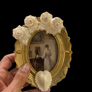 Pode incluir: Uma moldura oval dourada adornada com quatro conchas brancas e uma concha em forma de coração. A moldura contém uma impressão vintage de uma mulher em um vestido longo. A moldura está em um fundo preto.
