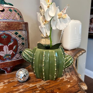 May include: A green ceramic cactus-shaped planter featuring small white dots along its vertical ribs. A white artificial orchid with yellow and pink centers is planted inside the cactus pot, which sits next to a small silver mirrored disco ball.