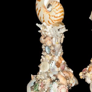 May include: A decorative candlestick covered in a dense mosaic of various seashells, including a large nautilus shell at the top. The structure features a mix of white, tan, brown, and iridescent abalone shell fragments arranged in a textured pattern.