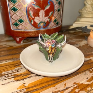 May include: A small green succulent-shaped planter holding a decorative bull skull ornament. The skull is encrusted with multicolored rhinestones, including clear, amber, pink, and blue crystals, and features prominent curved horns.
